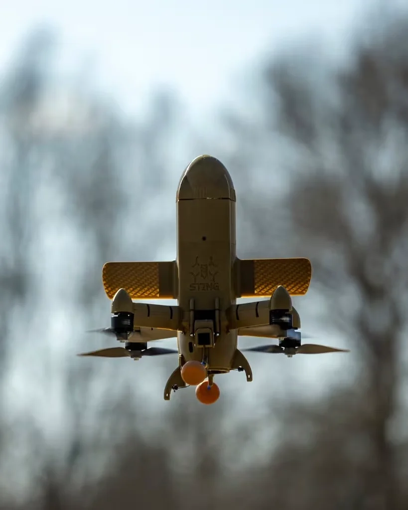Fixed-wing hybrid drone in flight, seen from below against bare winter trees.